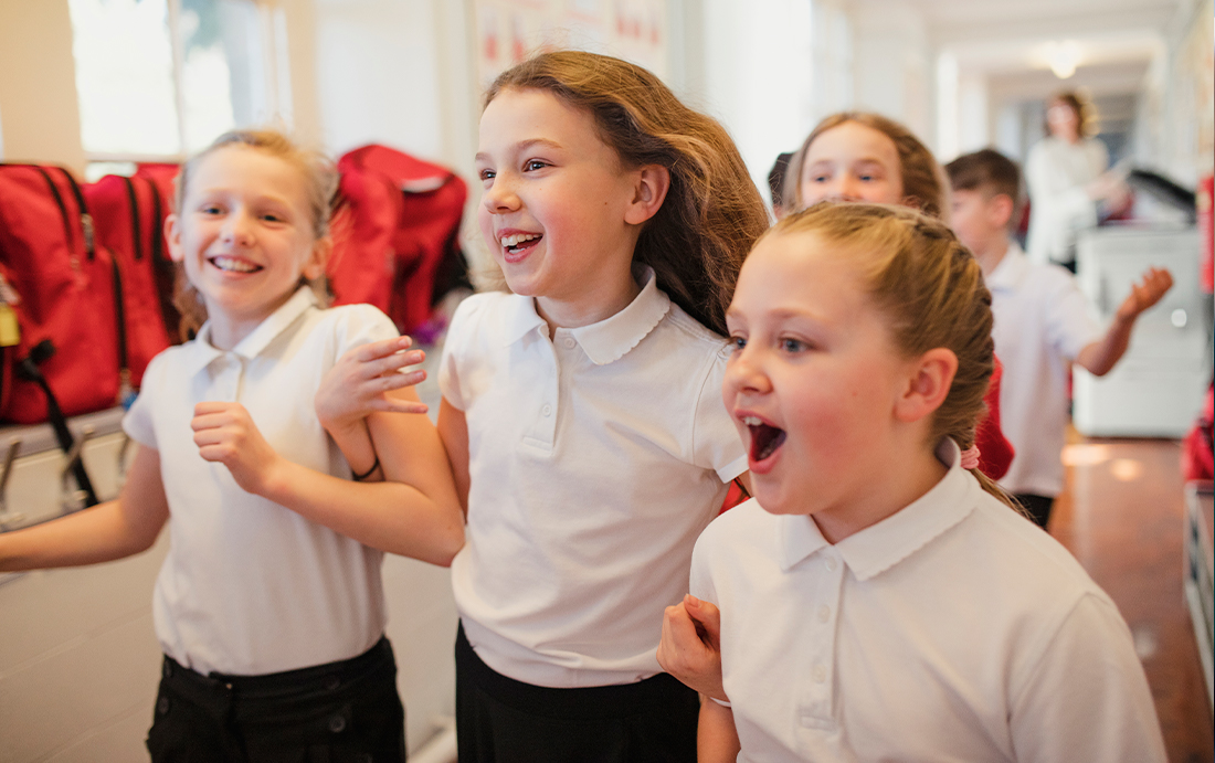 Three primary school-aged girls in uniform run smiling down a corridor